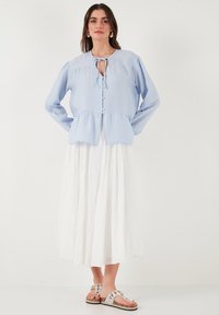 Blusa azzurro chiaro con colletto a cravatta, maniche arricciate e orlo a peplo, abbinata a una gonna bianca sopra il ginocchio e sandali a fantasia.