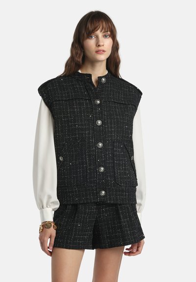 Femme portant un gilet sans manches texturé noir à boutons et un short assorti par-dessus un chemisier blanc à manches longues, debout devant un fond uni.