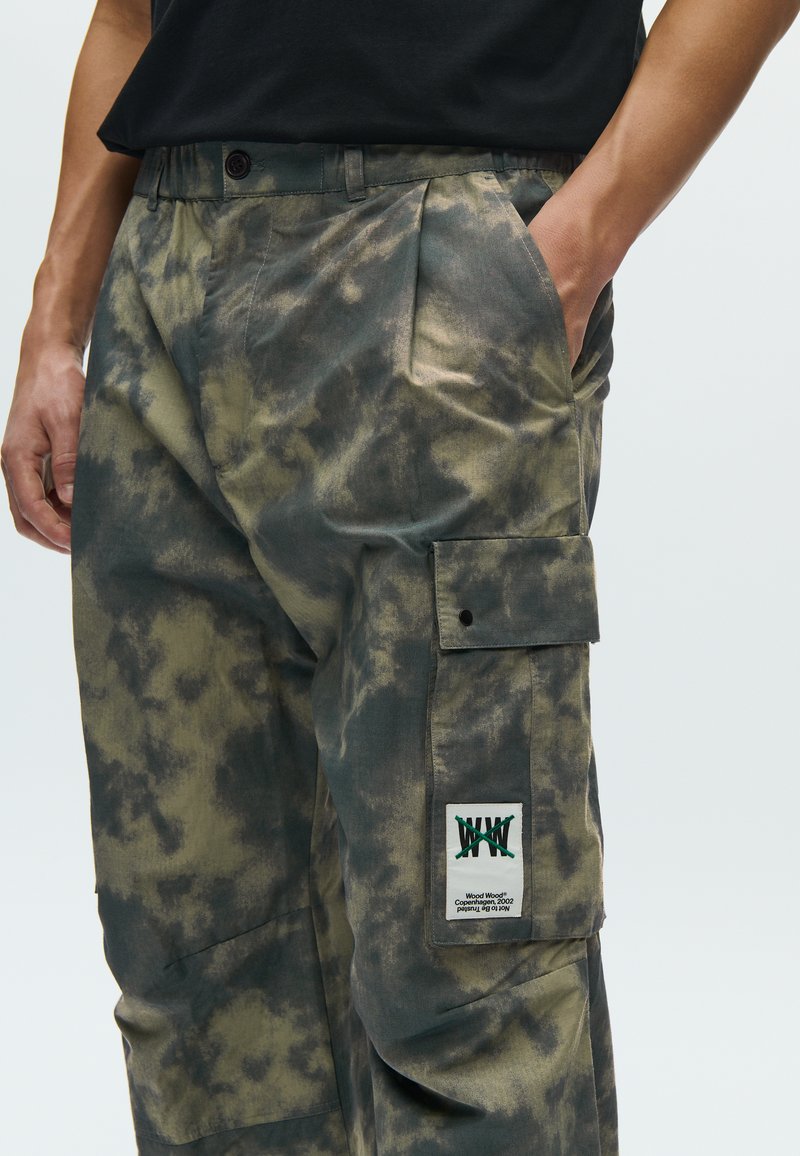 Pantalon cargo avec un motif camouflage vert, fabriqué en tissu doux. Comprend plusieurs poches, une fermeture à boutons, et une étiquette logo sur le côté.