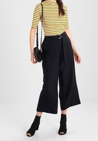 Pantalones de pierna ancha negros con cintura ceñida por un cinturón, combinados con una blusa de manga corta a rayas en amarillo y blanco. El modelo lleva un pequeño bolso negro.