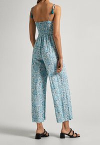 Modrý overal s květinovým vzorem, s elastickým pásem na zádech, širokými nohavicemi a dekorativními vázacími ramínky. Černé sandály na blokovém podpatku doplňují outfit.