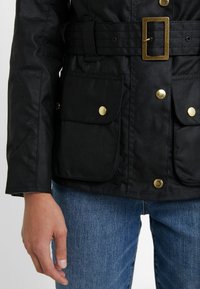 Schwarze Jacke aus glattem Stoff mit tailliertem Schnitt, goldenen Beschlägen und zwei großen Vordertaschen. Getragen über blauen Jeans.