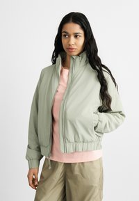 Νεαρή γυναίκα με μακριά σκούρα μαλλιά που φοράει ένα ανοιχτό πράσινο μπουφάν με φερμουάρ, ροζ πουκάμισο και μπεζ παντελόνι, στέκεται μπροστά σε ένα απλό λευκό φόντο.