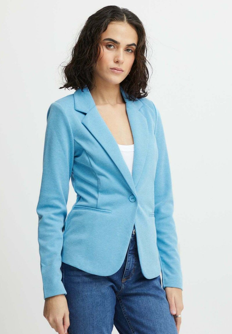 Blazer bleu clair avec une finition texturée, fermeture à un bouton, revers crantés et deux poches avant. Porté sur un haut blanc.