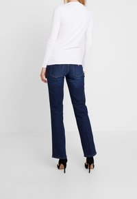 Camicia bianca a maniche lunghe e jeans dritti blu scuro. I jeans presentano tasche posteriori con cuciture sottili e sono abbinati a tacchi neri.