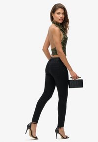 Zwarte high-waisted jeans in combinatie met een rugloze groene haltertop met pailletten. Houdt een gestructureerde zwarte handtas vast en draagt zwarte stilettohakken.