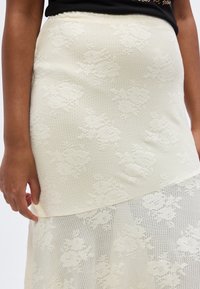 Κοντινό πλάνο ενός ατόμου που φοράει φούστα από κρεμ ύφασμα με υφή και λουλουδάτο δαντέλα, επενδεδυμένη με διάφανο δαντελένιο ύφασμα, με το ένα χέρι κοντά στο τελείωμα της φούστας.