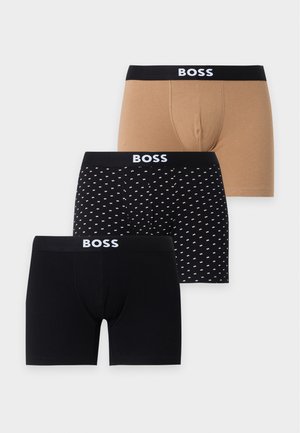 Muž bez košile s krátkými tmavými vlasy, který nosí béžové boxerky s černým páskem označeným BOSS, stojí bosý na světlém pozadí.