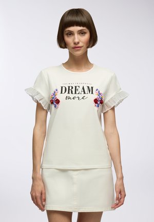 T-shirt bianco a maniche corte con bordi arricciati, ricamo floreale in rosso e blu, e la scritta "DREAM more" stampata in bold nero. Texture liscia.