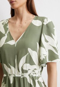 Abito verde con scollatura a V, maniche corte e un motivo floreale bianco. Il tessuto appare liscio, con una vestibilità comoda e vita arricciata.