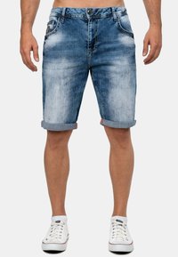 Shorts en denim bleu clair, avec un effet délavé, des ourlets retroussés et des poches avant avec rivets. Portés avec des baskets blanches.