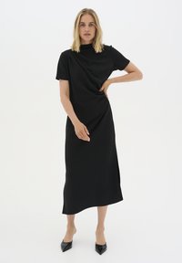 Black midi dress with short sleeves, draped design, high neckline, side slit, made from soft fabric. Paired with black pointed-toe heels.