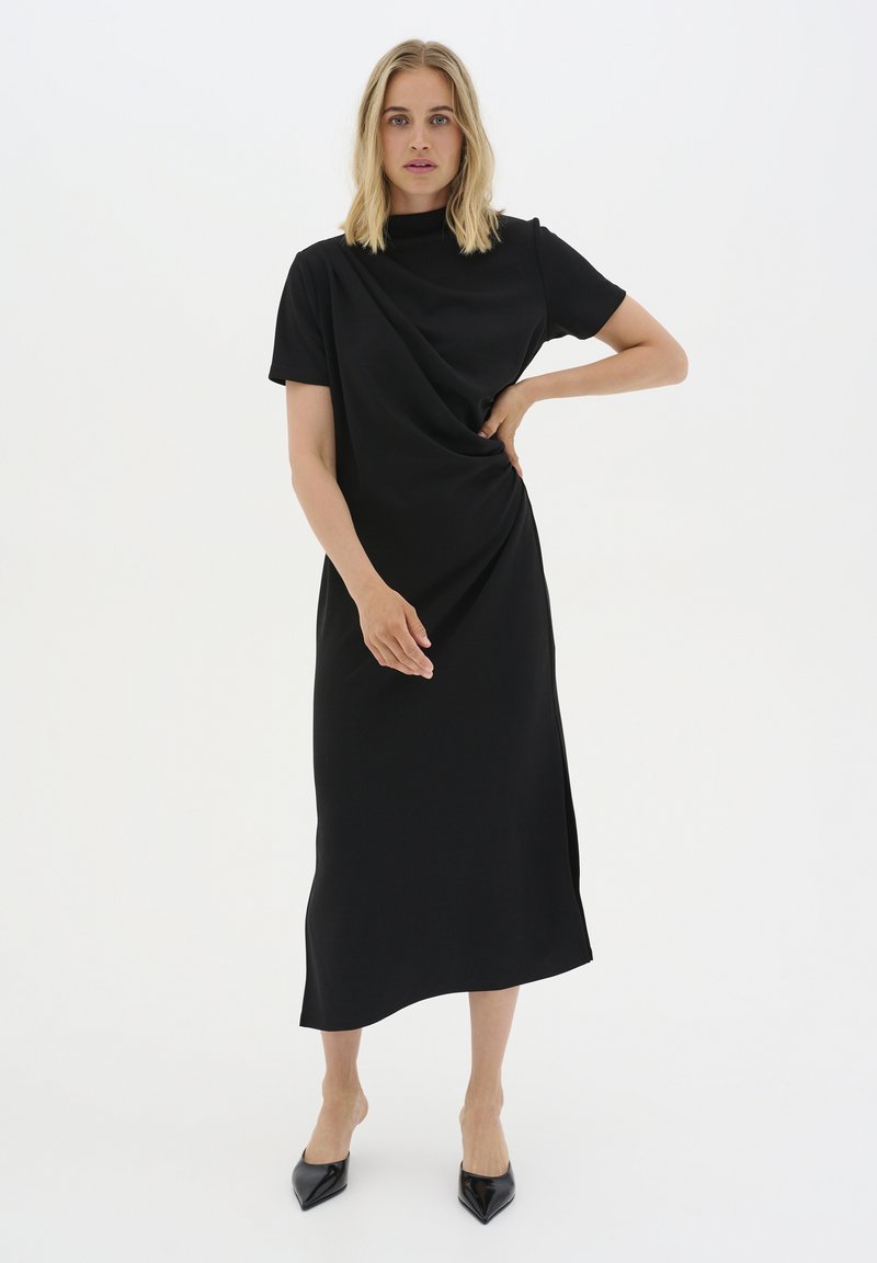 Black midi dress with short sleeves, draped design, high neckline, side slit, made from soft fabric. Paired with black pointed-toe heels.