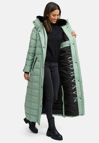 Lang mintgrøn pufferjakke med kviltet design, sort faux fur-krave, sort indvendig med branding, parret med sorte jeans og klodsede støvler.