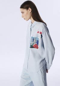 Camicia button-up di un leggero blu con una tasca a toppa decorata da ricami floreali in rosso, rosa e blu. Tessuto morbido con una vestibilità rilassata.