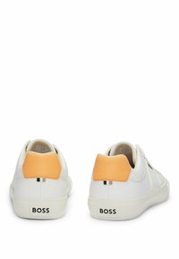 Zapatillas deportivas blancas con un acabado de cuero liso, que presentan un tirador texturizado en naranja en el talón y acentos negros en los laterales. La marca aparece en el talón.