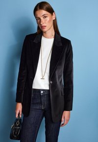 Blazer en velours noir avec revers crantés, fermeture à un bouton, associé à un haut blanc et un jean en denim foncé. Sac à main noir brillant en accent.