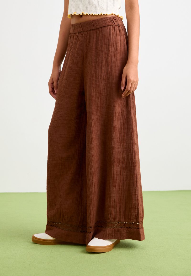 Pantaloni larghi marroni in tessuto rugoso, con una alta vita elastica e dettagli in pizzo sull'orlo, abbinati a scarpe chiare.