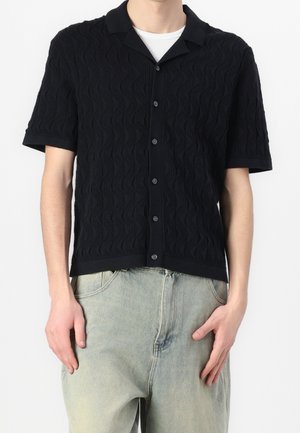 Camisa negra de manga corta con un patrón de ondas texturizado, con cuello y cierre de botones, llevada sobre vaqueros de mezclilla de color claro.