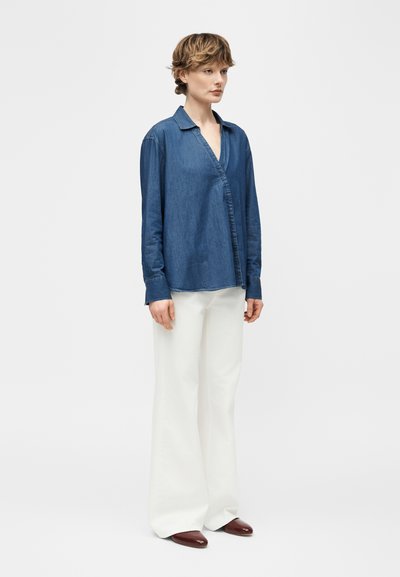 Γυναίκα που στέκεται, φοράει μπλε πουκάμισο τζιν με μακριά μανίκια, φαρδιά λευκή παντελόνα και καφέ παπούτσια μπροστά σε απλό φωτεινό φόντο.
