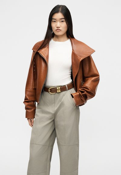 Jeune femme aux longs cheveux noirs portant une veste en cuir marron sur un haut blanc, un pantalon beige taille haute et une large ceinture marron.