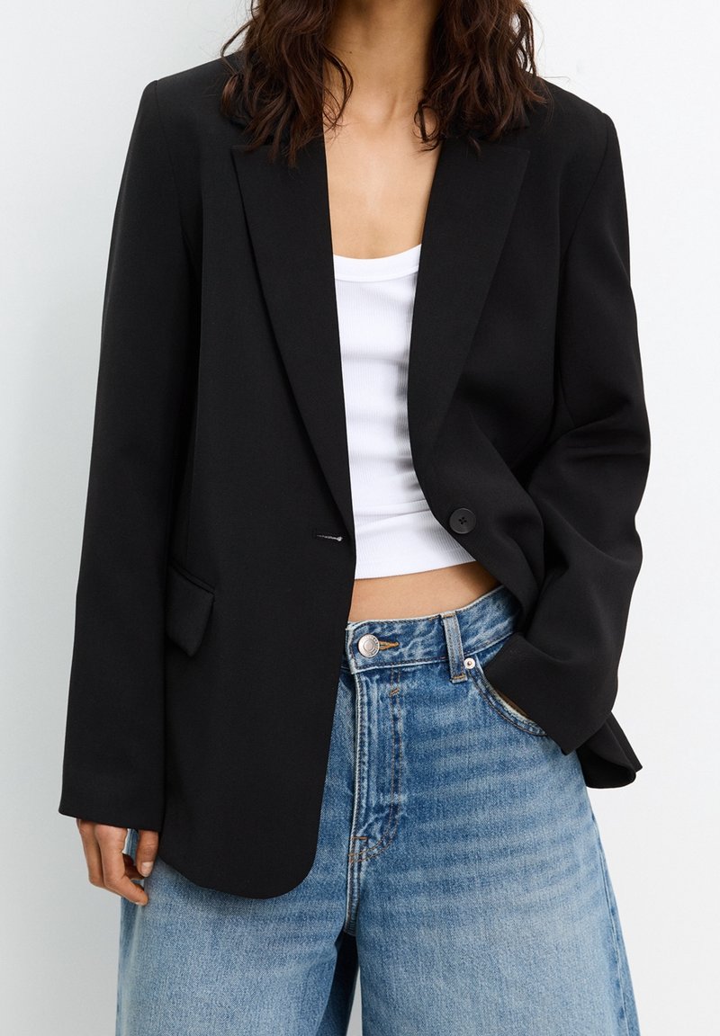 Blazer noir avec une fermeture à un seul bouton, des revers crantés et des poches, porté sur un débardeur blanc et un jean taille haute bleu.