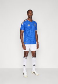 Plava nogometna majica s teksturiranim uzorkom, zlatnim detaljima, grbom talijanske zastave, bijele kratke hlače, bijele čarape s crnim prugama i crne kopačke.