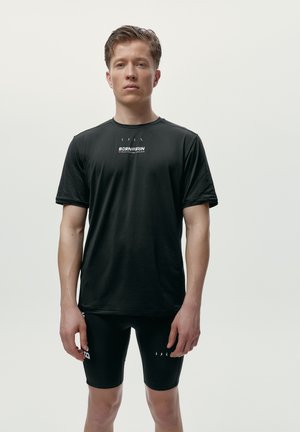 En ung mand iført en sort t-shirt med korte ærmer og sorte cykelshorts står mod en ensfarvet lys baggrund.