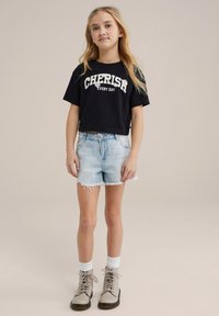 Schwarzes, kurzes T-Shirt mit dem Aufdruck "JEDEN TAG SCHÄTZEN", hellblaue Denim-Shorts mit fransenförmigen Kanten und hellbraune Schnürstiefel mit weißen Socken.