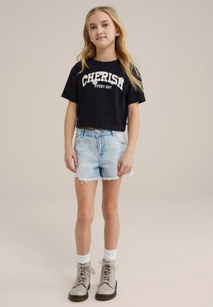 T-shirt noire raccourcie avec le texte "CHÉRIR CHAQUE JOUR", short en denim bleu clair avec des bords effilochés et bottes marron lacées avec des chaussettes blanches.