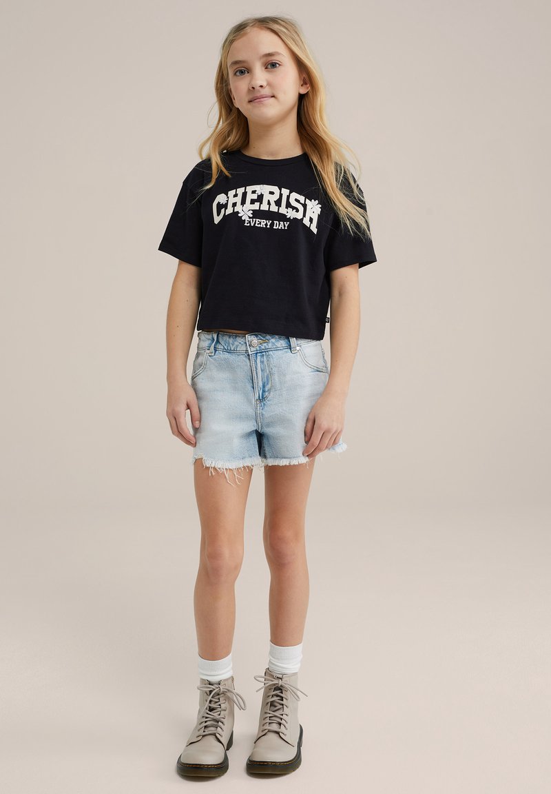Schwarzes, kurzes T-Shirt mit dem Aufdruck "JEDEN TAG SCHÄTZEN", hellblaue Denim-Shorts mit fransenförmigen Kanten und hellbraune Schnürstiefel mit weißen Socken.