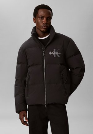 Νεαρός άνδρας που φοράει μαύρο μπουφάν Calvin Klein με ψηλό γιακά, στέκεται μπροστά σε μια απλή γκρι φόντο.