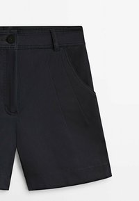 Shorts noirs ajustés avec plis à l'avant, poches latérales, fermeture par boutons et passants pour ceinture sur fond blanc.