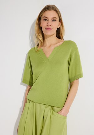 Vrouw met blond haar, gekleed in een lichtgroene kortemouw V-hals trui en bijpassende rok met grote zakken, staand tegen een witte achtergrond.