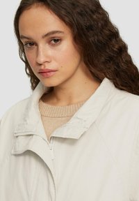 Licht beige zip-up jack met een hoge kraag, zachte katoenstof en een gestructureerde gebreide trui eronder.