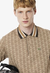 Barna mintás pólóing, gallérral és csíkozott részletekkel. Egy zöld logó található a gombos slicc felett.