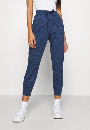 Kobieta w niebieskich, wzorzystych spodniach dresowych na sznurek, białych skarpetkach, białych trampkach oraz białym topie bez rękawów na jednolitym tle.