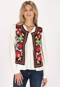 Schwarze ärmellose Weste mit mehrfarbiger Blumenstickerei in Rot, Grün und Weiß, verziert mit roten und schwarzen Details. Getragen über einem weißen Oberteil.