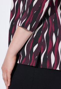 Kortärmad topp med ett mönster av mörkröd, svart och krämfärgad våg. Tyget är slätt med sidospalter och subtila sömnadsdetaljer.