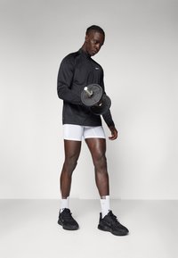 Svart atletisk jacka, vita shorts, håller i ett svart viktskott. Svarta sneakers med vita detaljer; fokuserad min, muskulös kropp.