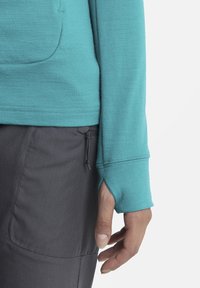 Camicia a maniche lunghe turchese con fori per il pollice, abbinata a pantaloni grigi con cordoncino. Tessuto morbido e leggero con una superficie liscia.