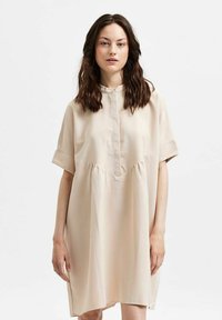 Kort beige kjole med kragehals, korte ermer og innsamlet midje. Laget av lett stoff med en glatt tekstur.