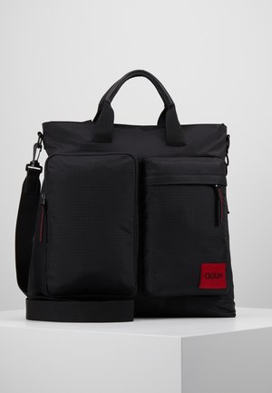 Musta suorakulmainen olkalaukku, jossa on kaksi etupuolen vetoketjullista taskua, yläkahvat, säädettävä olkahihna ja punainen Hugo-logo patch alakulman taskussa.