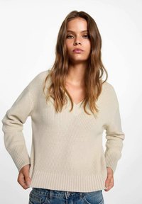 Beige stickad tröja med V-urringning och ribbad nederkant, med långa ärmar och avslappnad passform, bärs tillsammans med blå jeans.