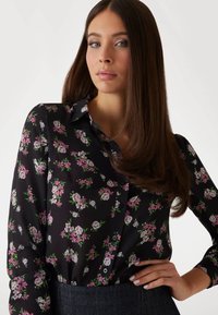 Blusa nera floreale con fiori rosa e bianchi, bottoni sul davanti, maniche lunghe, tessuto morbido e silhouette adatta.