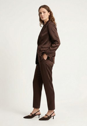 Vrouw met golvend haar die een bruine satijnen pak draagt met blazer en taps toelopende broek, gecombineerd met bruine slingback kittenheels, zijwaarts staand.