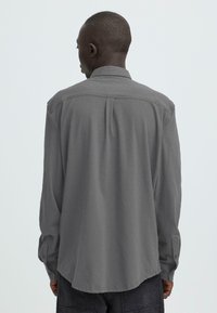 Chemise grise à manches longues avec col à boutons, coupe décontractée et poignets à boutons ajustables. Détail discret de plis à l'arrière.