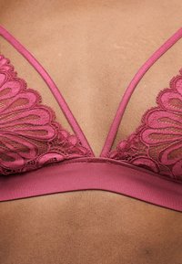 Κοντινό πλάνο ροζ δαντελένιου σουτιέν bralette με λεπτές τιράντες, φορεμένο πάνω σε δέρμα, με βαθύ ντεκολτέ σε σχήμα V και διακοσμητικά λουλουδάτα μοτίβα από δαντέλα.