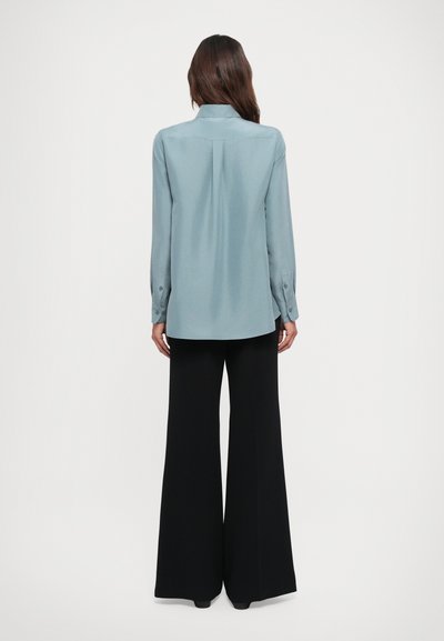 Light blue long-sleeve blouse with a collar, featuring a central pleat and buttoned cuffs, paired with black wide-leg trousers.