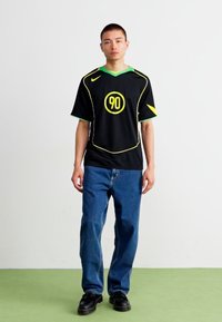 Svart T-skjorte med korte ermer og grønne detaljer samt gule kanter, med en sirkulær gul grafikk med '90' i sentrum, kombinert med blå jeans.